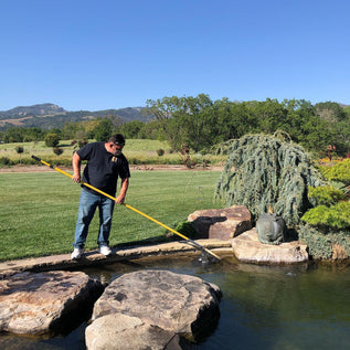 Person cleaning a pond with a long tool in a garden setting